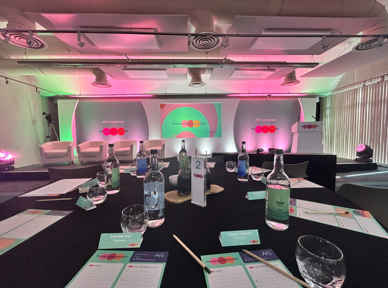 A conference room setup featuring a round table with notepads and pens. In the background, there is a colorful stage with lighting, equipped with a large screen and chairs arranged for a presentation.