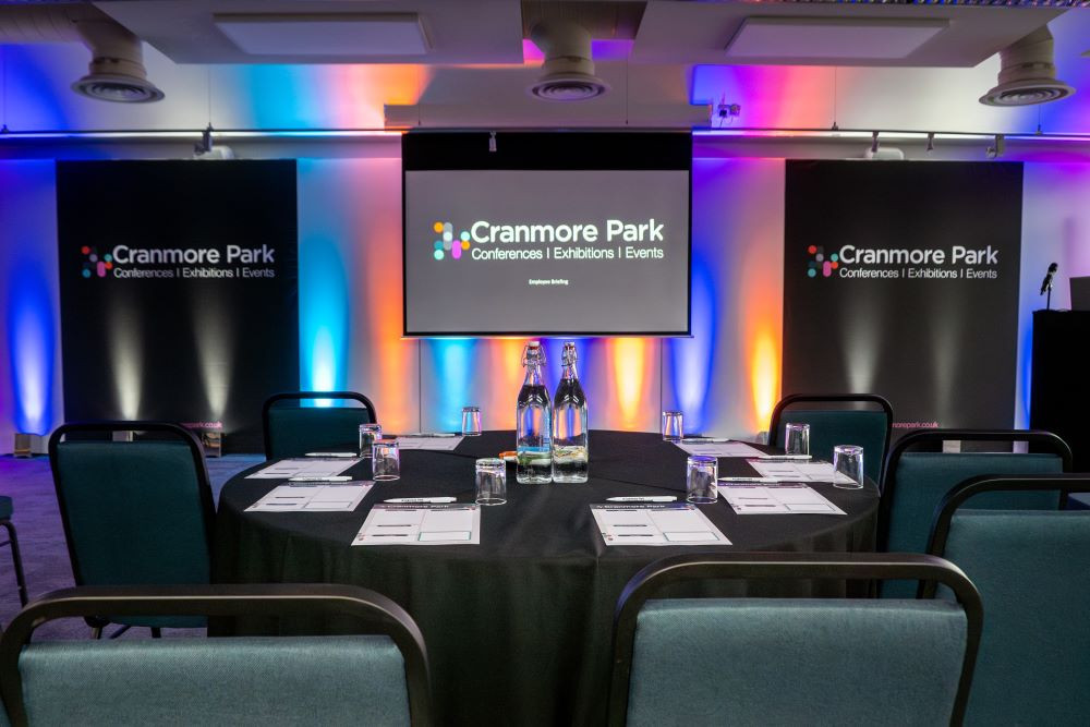 A conference setup with a round table covered in a black cloth, featuring bottles of water and glasses. Colorful ambient lighting enhances the space, with banners displaying 