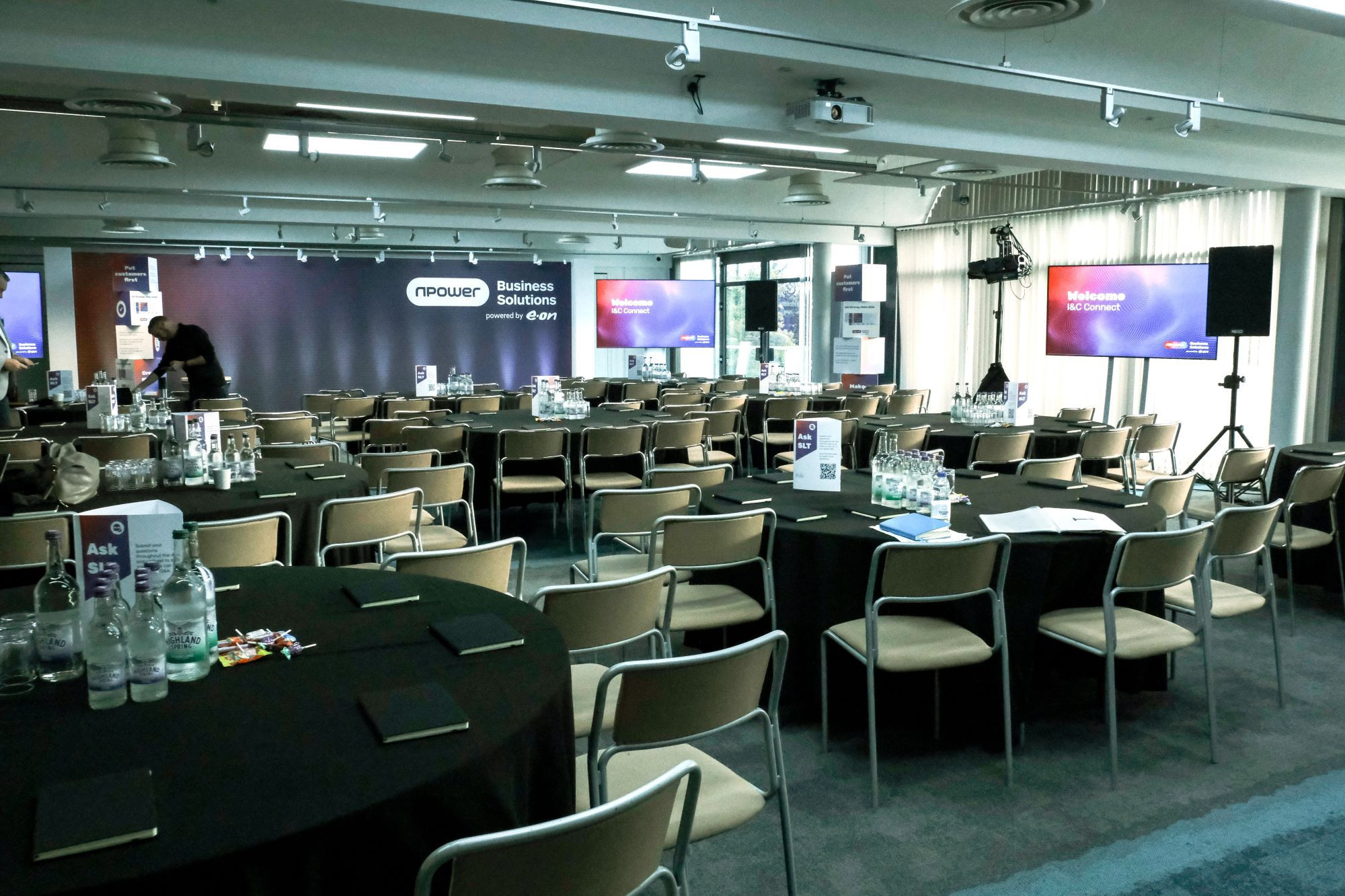 A conference room set up for an event, featuring rows of round tables with black tablecloths. Water bottles and notepads are on each table. A stage is in the background with large screens displaying a welcome message. The room is well-lit and has a modern design.