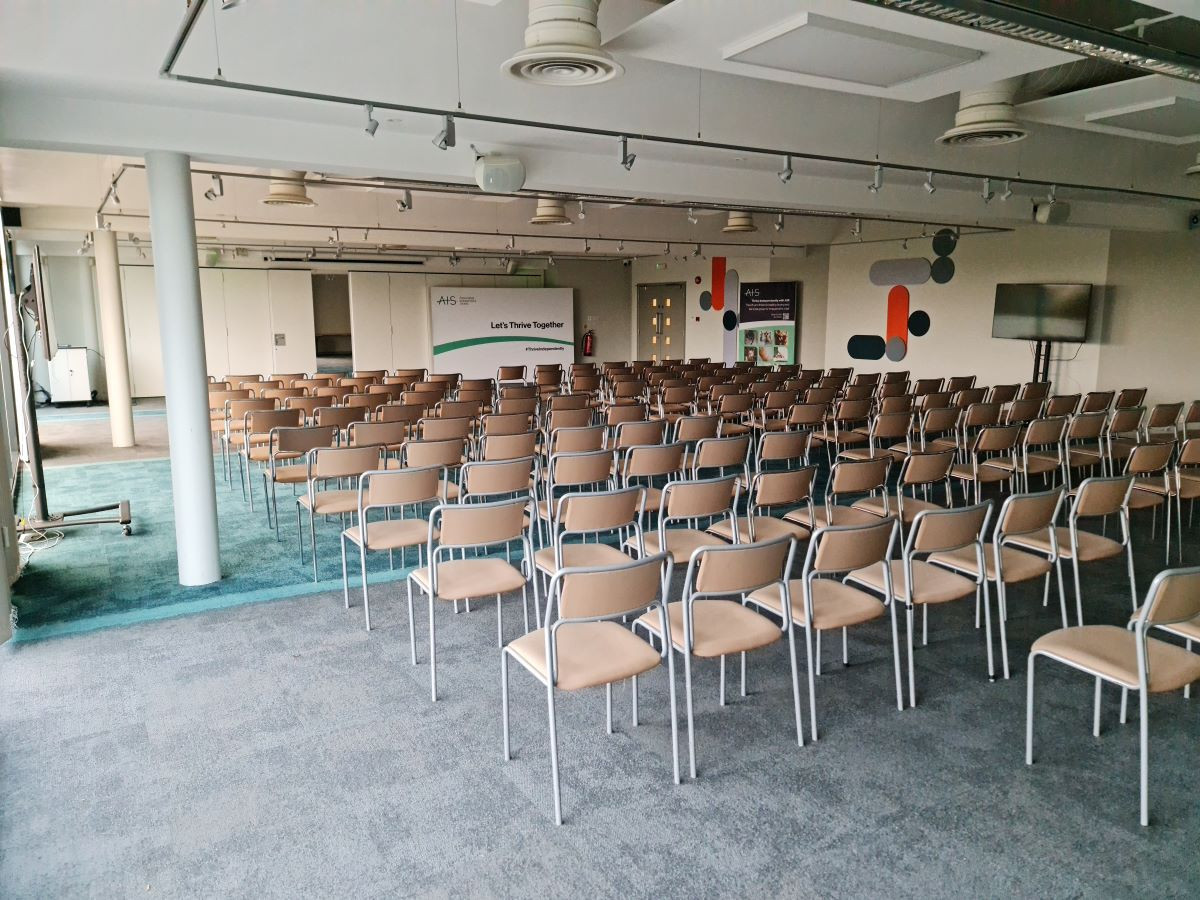 A spacious conference room with a row of beige chairs arranged in neat rows. The floor is carpeted in a muted color, and there are modern decor elements on the walls. A large screen is set up at the front of the room, and a banner in the background displays the words 