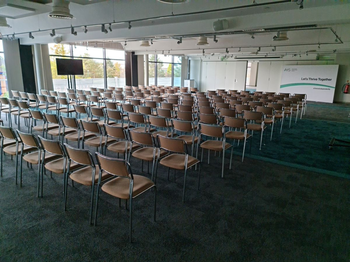 A spacious conference room with rows of empty metal and plastic chairs facing the front. Large windows provide natural light and a view of greenery outside. A screen is set up at the front, displaying the text 