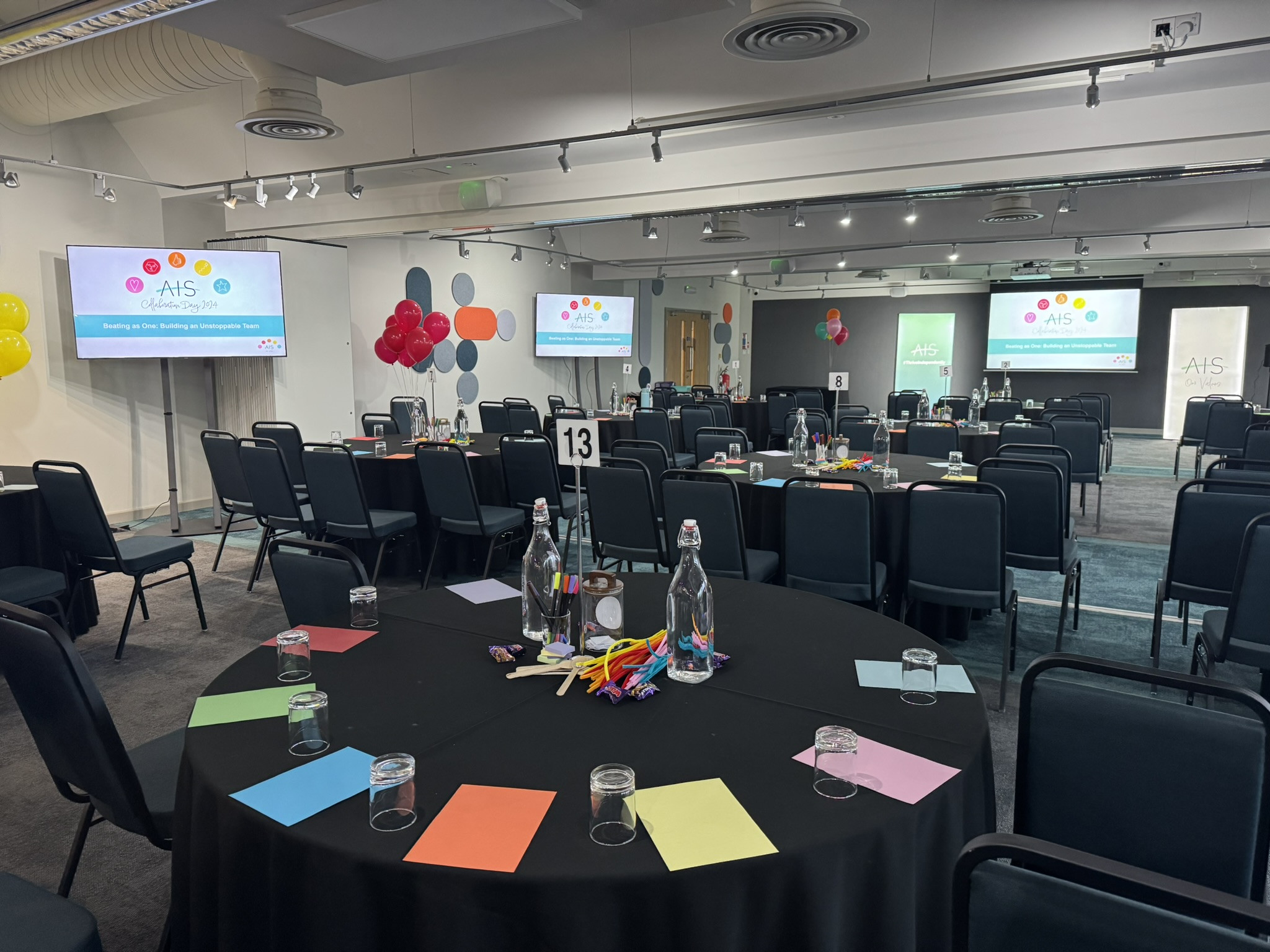 A conference room set up for an event, featuring several round tables with colorful place cards, water bottles, and stationery. Two large screens display the event logo, and balloons are decoratively arranged around the room.