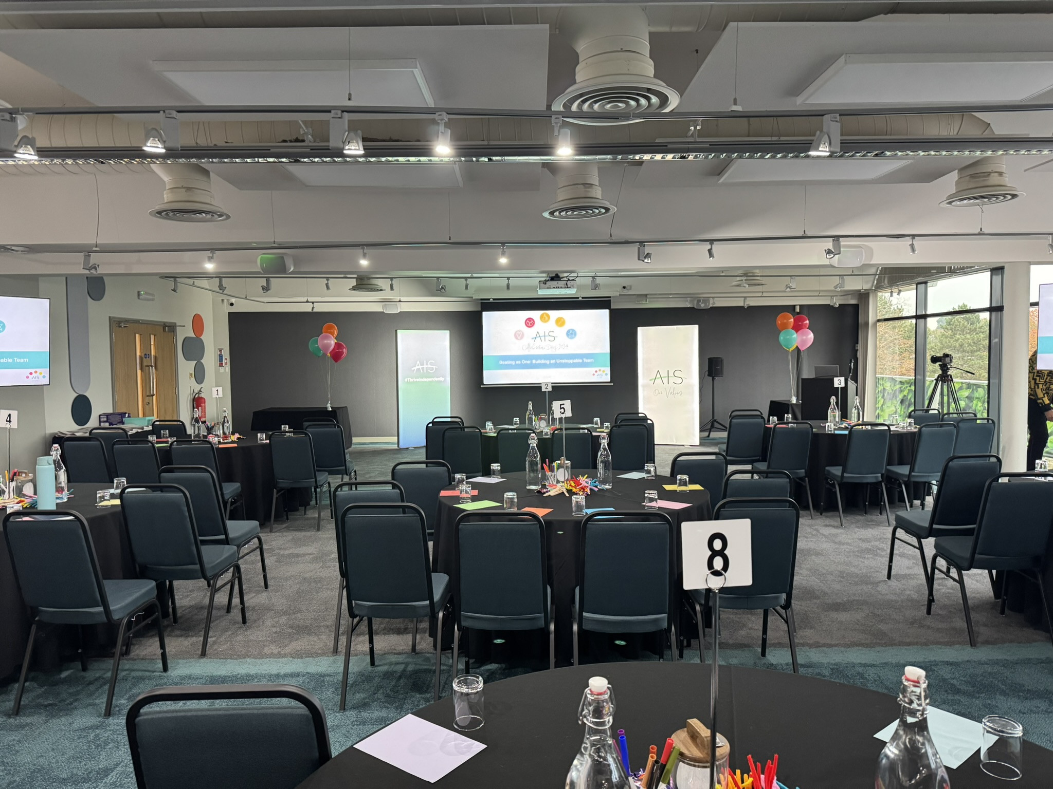 A large conference room set up for an event, with round tables covered in black cloths. Each table has water bottles, colorful pens, and name cards. Balloons decorate the space, and two large screens display presentation slides. The room is well-lit with windows showing greenery outside.