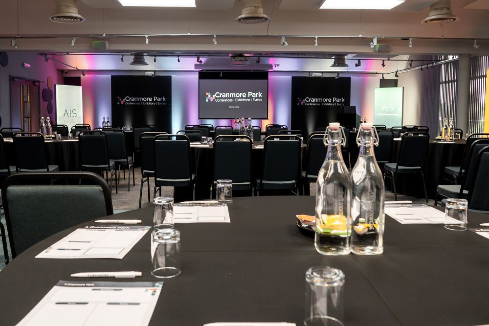 A conference room with round tables covered in black tablecloths, equipped with water bottles and glasses. Two screens at the front display the 