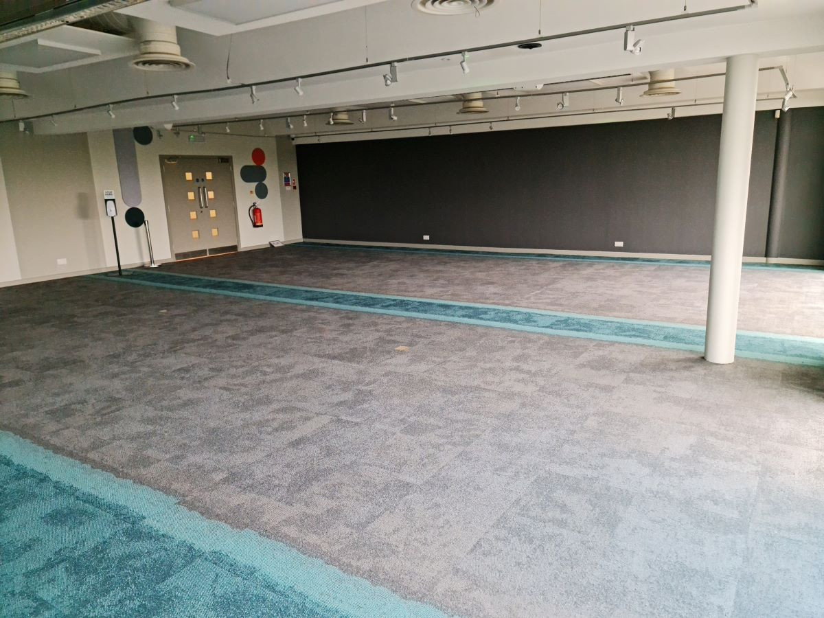 A spacious, empty interior room with gray carpet featuring turquoise accents. The walls are mostly dark, with a few colorful circular decorations. Bright overhead lights illuminate the area.