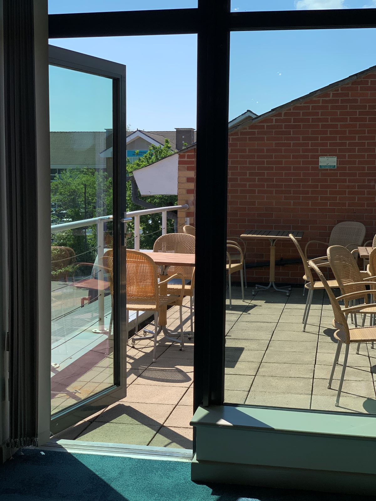 A sunny balcony with several wicker chairs and tables, viewed through an open glass door. The setting features a mix of brick and modern architecture in the background.
