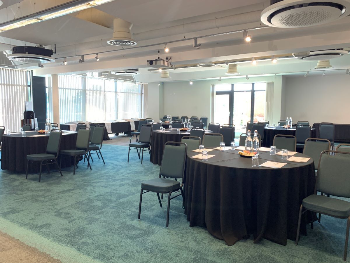 A spacious conference room set up with round tables covered in black tablecloths. Each table has water pitchers and glasses. The room features large windows with plenty of natural light and modern lighting fixtures on the ceiling.