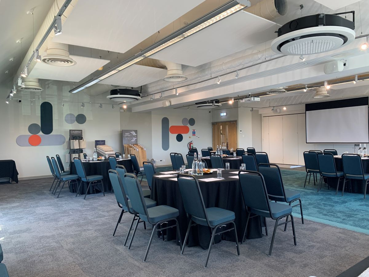 A conference room set up with multiple rows of dark blue chairs around tables covered in black cloth. The walls feature abstract designs in muted colors, and a projector screen is visible at the front of the room. Natural light illuminates the space.