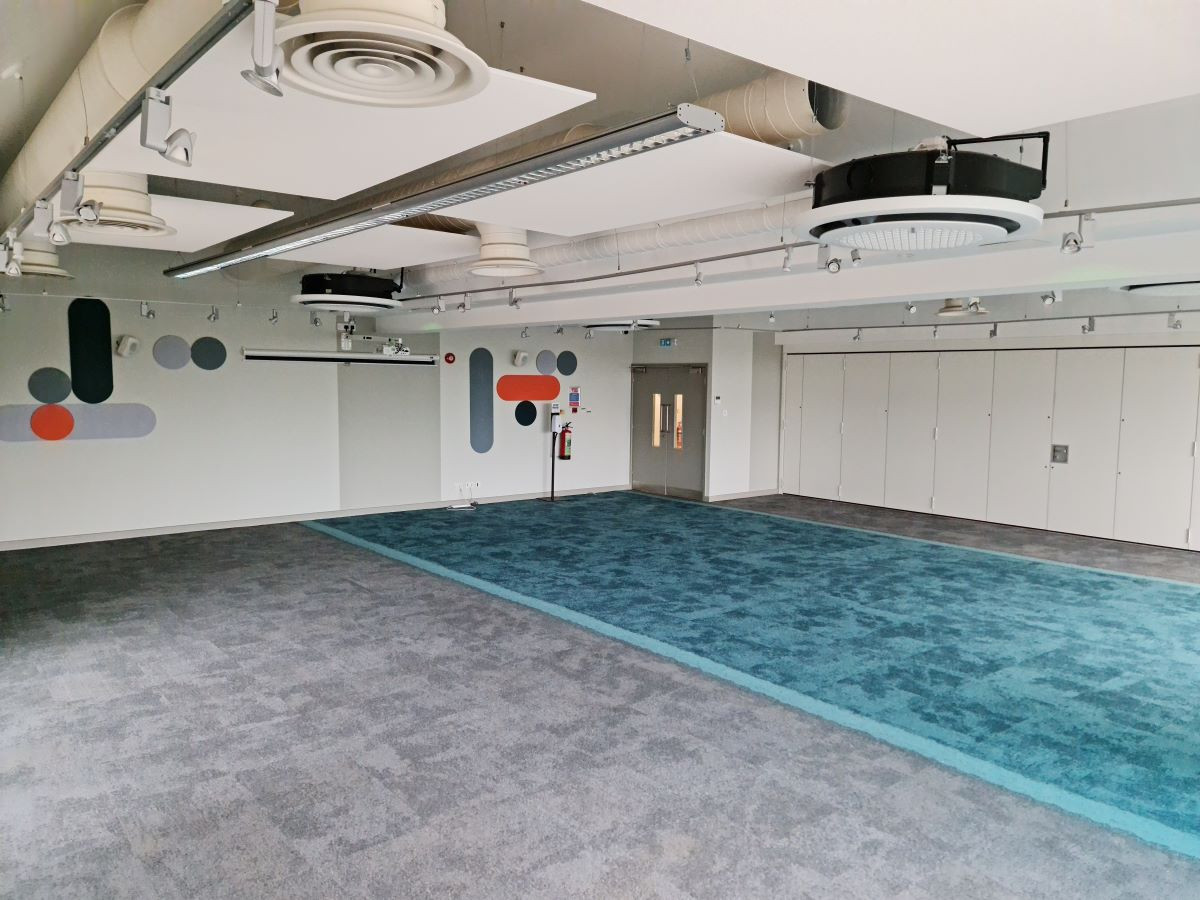 A spacious, modern room with gray carpet and a large teal carpet area. Walls feature abstract designs in orange, blue, and gray. There are ceiling-mounted lights and a projector system visible. Storage cabinets line one side of the room.