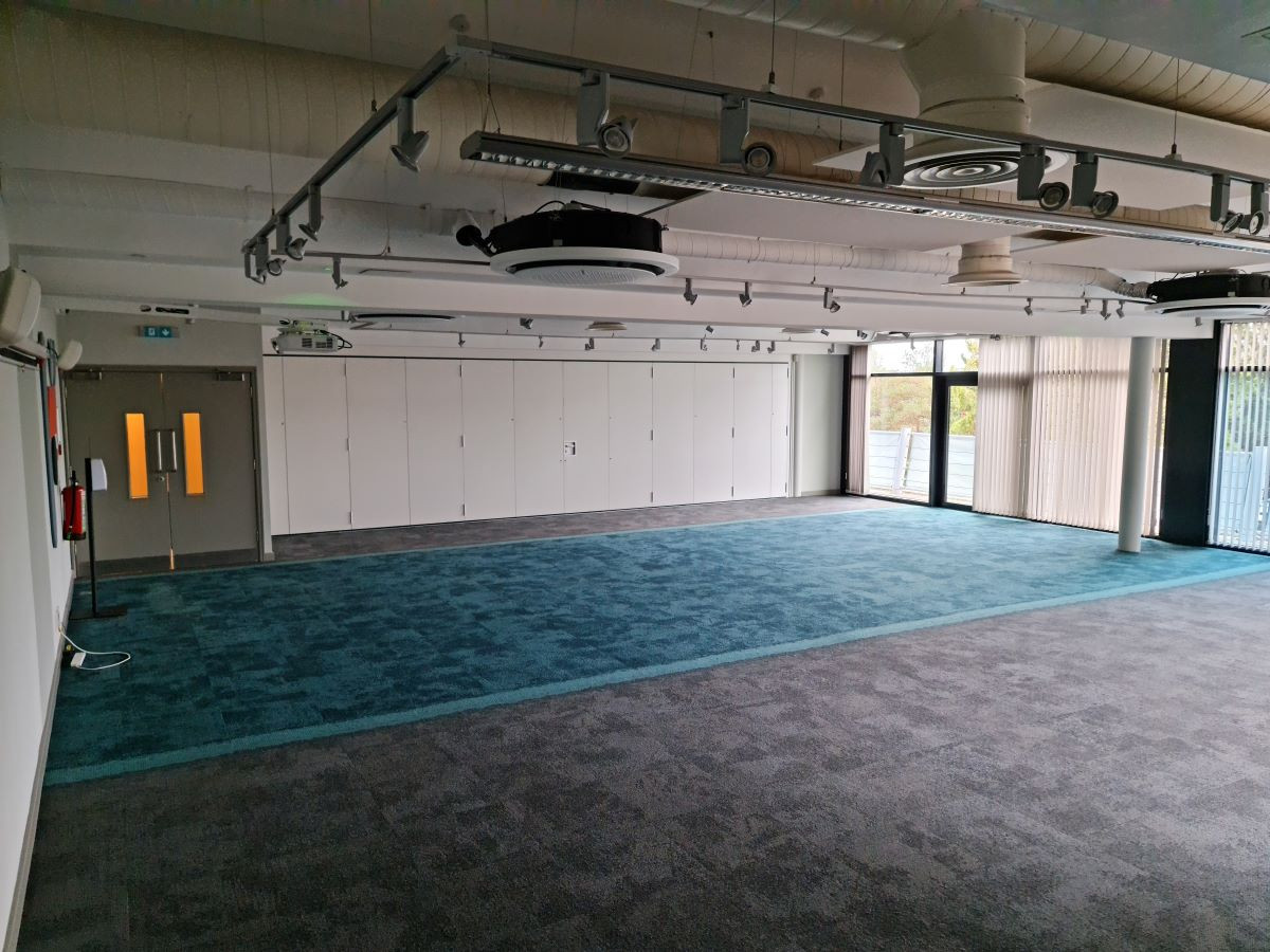 Empty meeting room with grey and teal carpeting, large windows allowing natural light, and storage cabinets along one wall. Ceiling features exposed ducts and lighting fixtures.