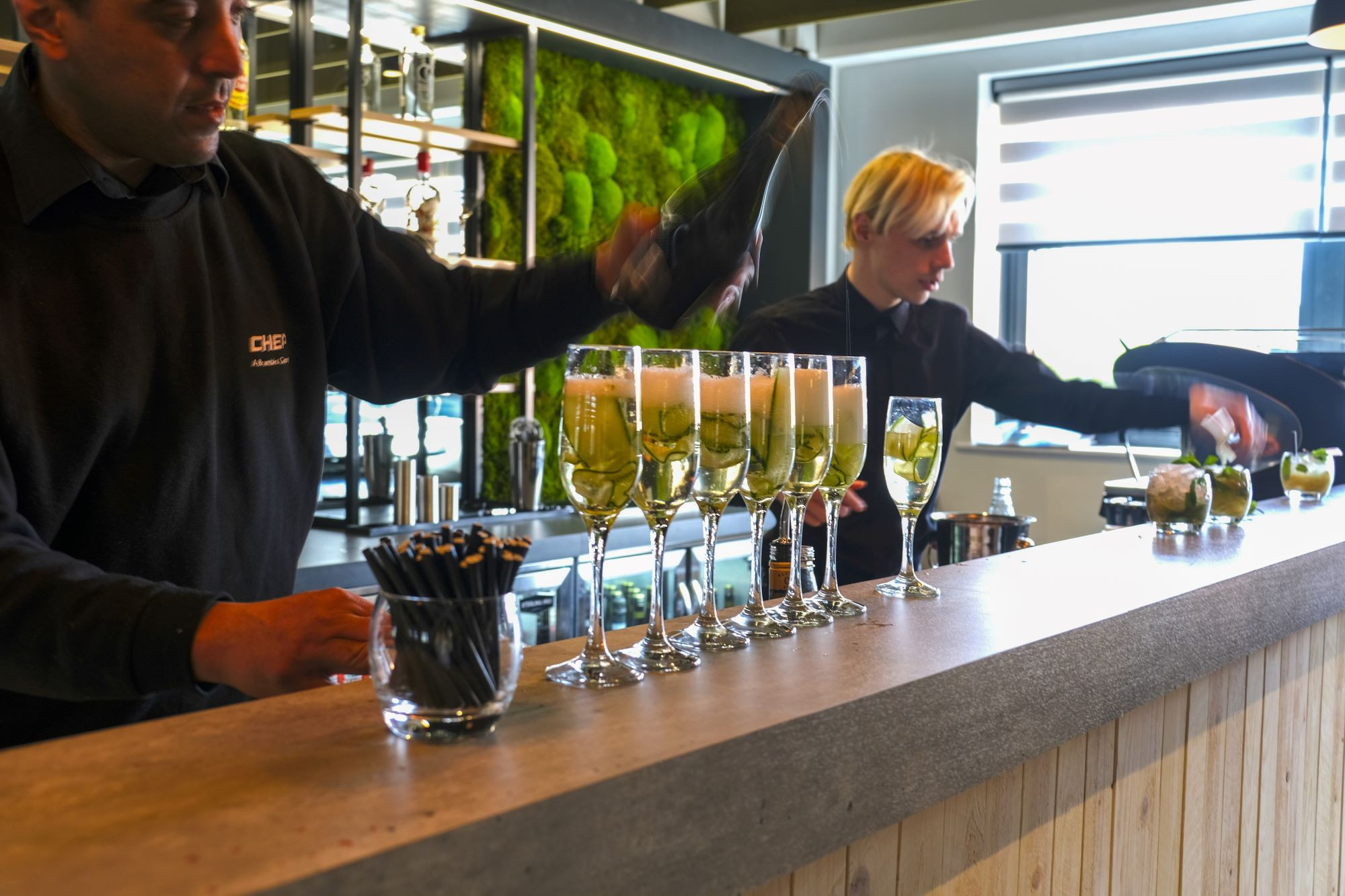 A bartender pours champagne into glasses lined up on a bar counter, while another staff member prepares drinks in the background. The bar features green moss decor, adding a natural touch to the modern setting.