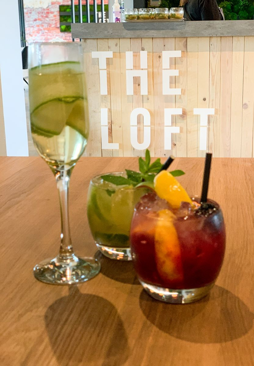 Three cocktails are arranged on a wooden table. On the left, a tall glass filled with a green drink and cucumber slices. In the center, a short glass contains a green cocktail, garnished with mint leaves. On the right, a short glass holds a dark red drink with an orange slice and a cherry on top. Behind them, the words 